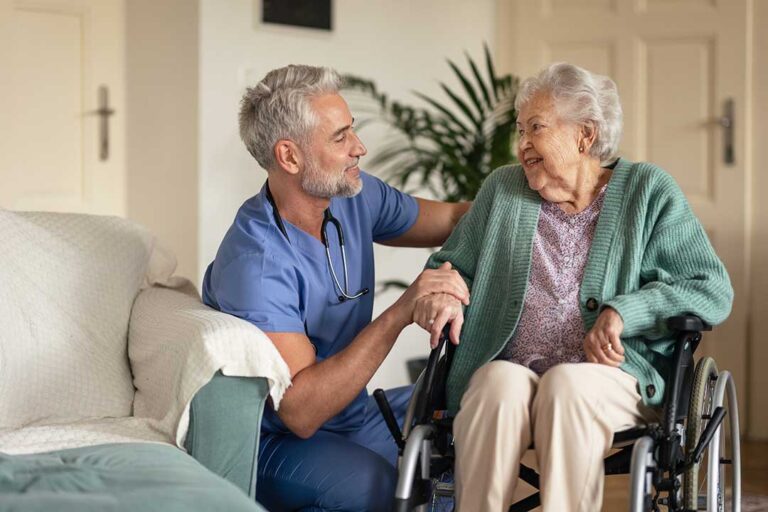 An elderly woman chatting with a caregiver while sitting at a small dining table in a sunny residential room.